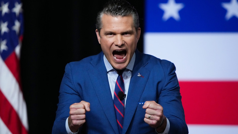 Pete Hegseth balling both his fists as he screams in a blue suit in front of an American flag