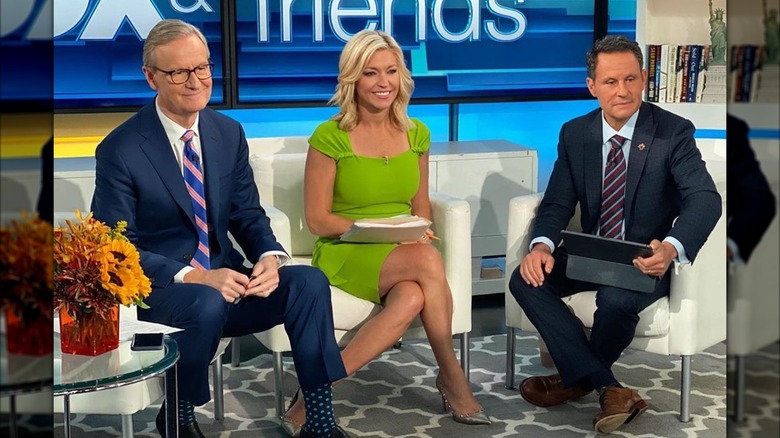 Ainsley Earhardt looking off with a smile beside two men, all seated.