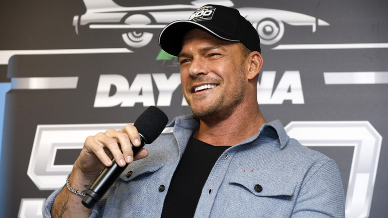 Alan Ritchson smiling and holding a microphone and wearing a Daytona 500 hat.