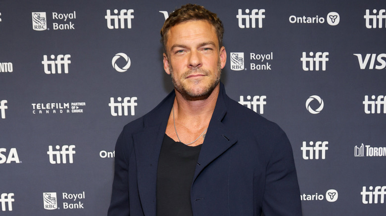 Alan Ritchson posing in a black shirt and dark blue blazer.