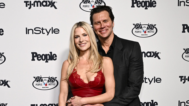Ali Larter posing in a red dress while her husband Hayes MacArthur hugs her from behind in a black suit