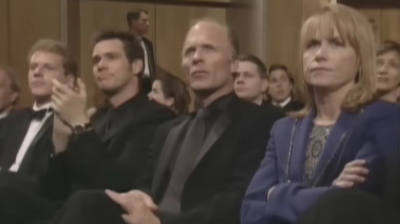 Ed Harris and Amy Madigan in the audience at the Oscars with their arms crossed while others, including Jim Carrey, applaud