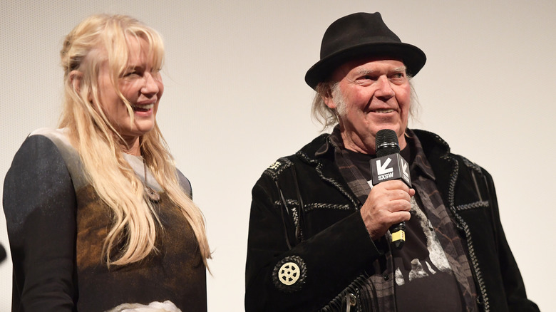 Daryl Hannah smiling in black with Neil Young in a hat holding a mic