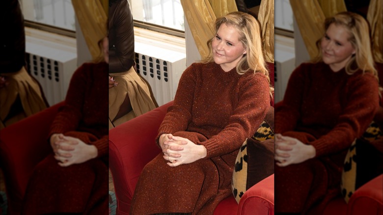 Amy Schumer sitting on a couch wearing a dark orange sweater and skirt