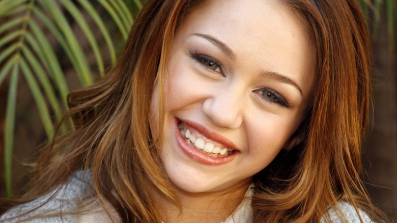 A young Miley Cyrus smiling in front of a palm frond
