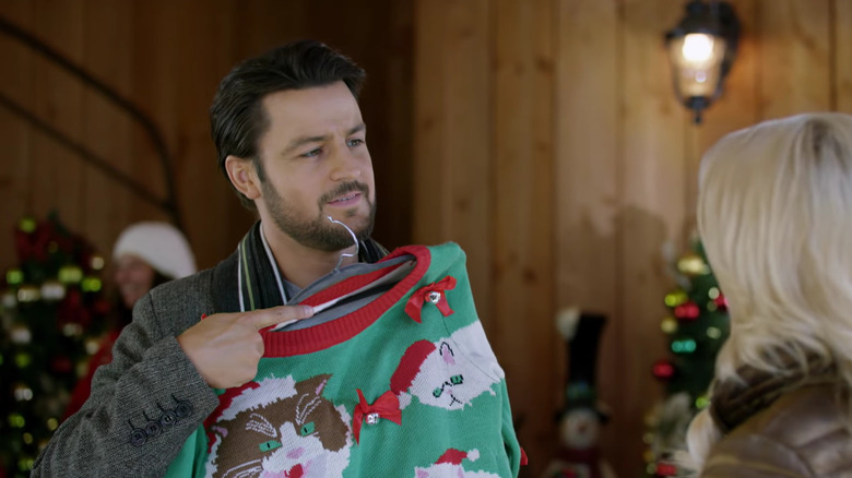 Tyler Hynes holding up a Christmas sweater while a woman watches