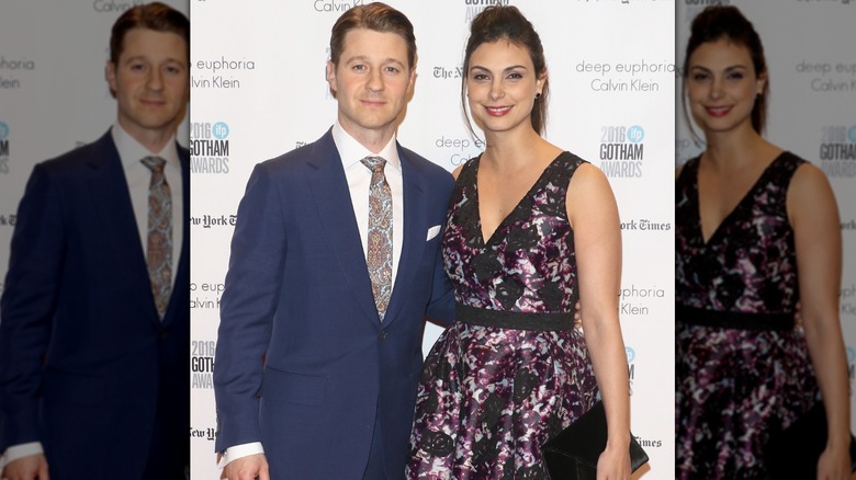 Ben McKenzie wears a dark blue blazer and poses with his arm around Morena Baccarin, who wears a purple floral-print dress.