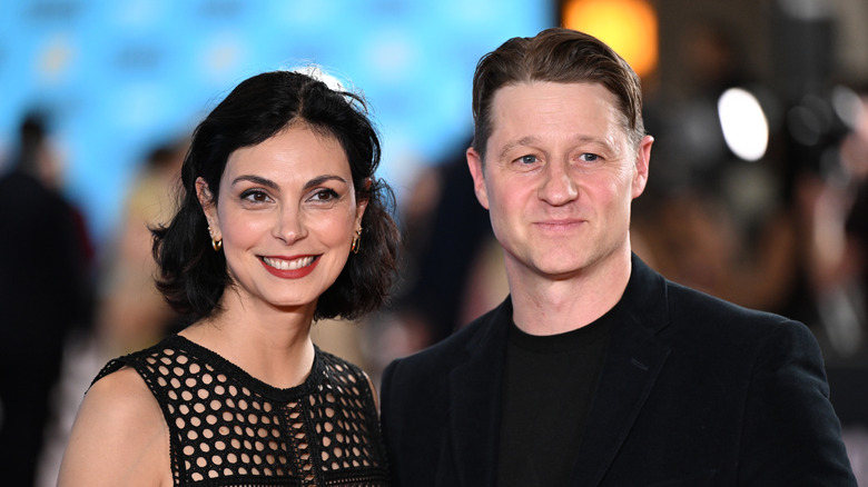 Morena Baccarin wears a black mesh dress and poses next to Ben McKenzie, who wears a black blazer and shirt.