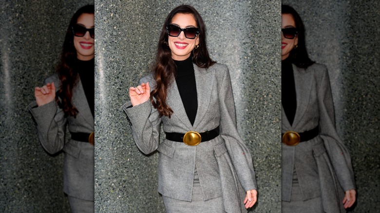 Anne Hathaway waves and smiles in a belted gray plaid coat and skirt.
