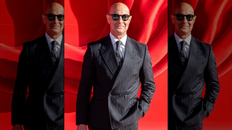 Stanley Tucci posing with a hand in his pocket in a black pinstripe suit and sunglasses.
