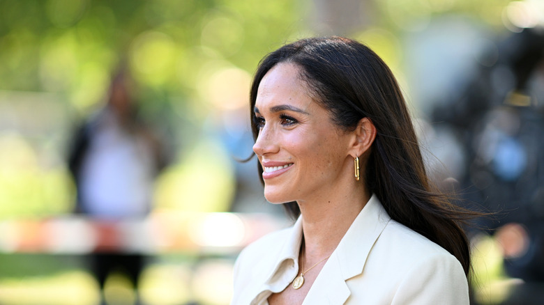 Smiling Meghan Markle wearing a white blazer with gold jewelry