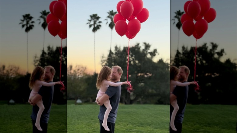 Princess Lilibet, dressed in ballet clothes, holding a bunch of red balloons as Prince Harry holds her in his arms