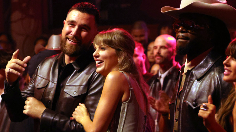 Taylor Swift at an event with others holding on to Travis Kelce's arm