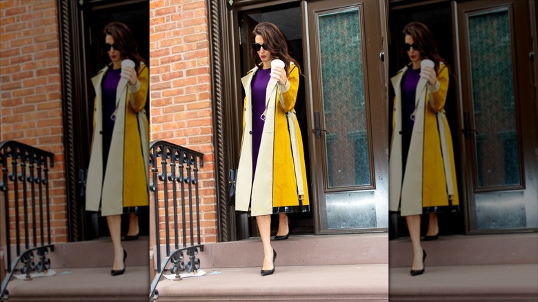 Amal Clooney in a yellow jacket walking down stone steps with a coffee in hand.