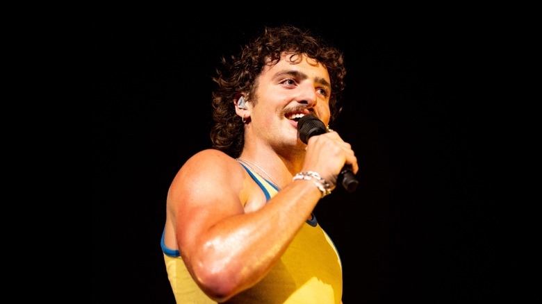 Benson Boone in a yellow tanktop holds a microphone to his mouth on a black background