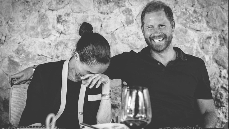 Meghan Markle laughing and Prince Harry smiling at dinner table in front of stone wall