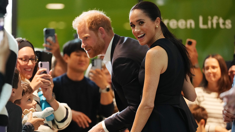 Meghan Markle smiling while Prince Harry greets a crowd