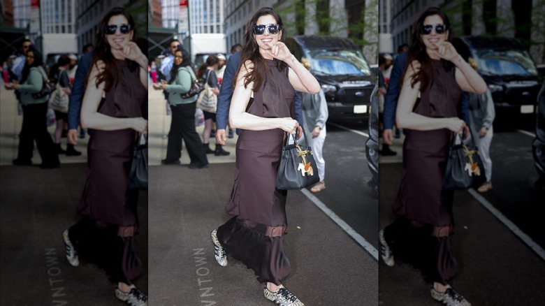 Anne Hathaway waving to camera in brown dress, sneakers, and sunglasses