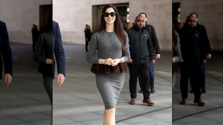 Anne Hathaway smiling while walking in a gray dress with brown peplum and sunglasses