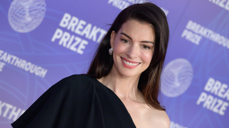 Anne Hathaway smiling while posing in a black one-shoulder dress