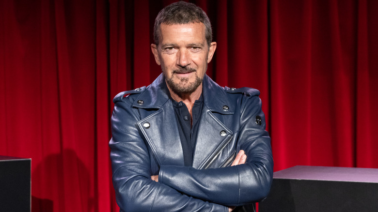 A smiling Antonio Banderas posing with his arms crossed in a black leather jacket with a black shirt underneath
