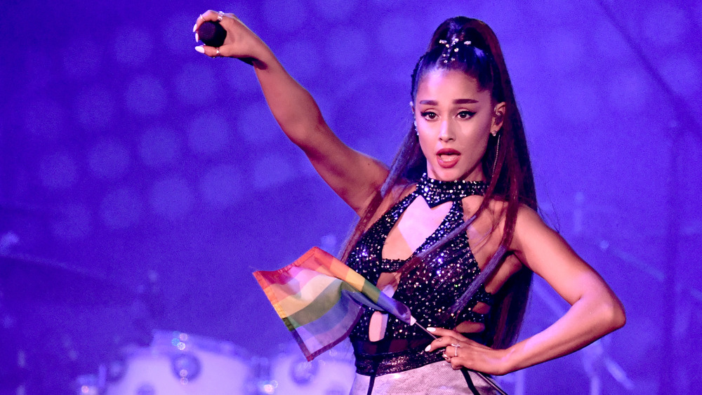 Ariana Grande holding a mic out to the audience, holding a rainbow flag, while singing