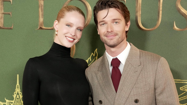 Abby Champion and Patrick Schwarzenegger smiling while walking a White Lotus red carpet.