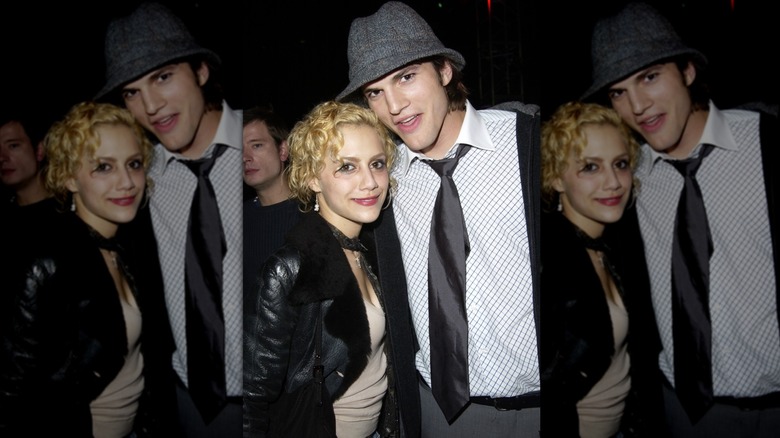 Brittany Murphy in a beige top and black jacket with crimped blond hair, smiling next to Ashton Kutcher in a gray hat and matching tie