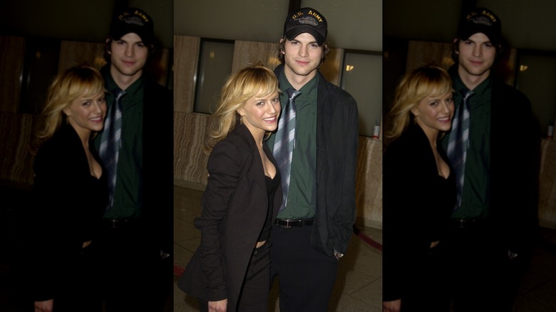 Brittany Murphy smiling and wearing all black next to smiling Ashton Kutcher, in a green shirt with a baseball cap on his head