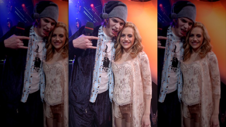 Ashton Kutcher making a "rock on" hand sign in a mismatched outfit with a stocking cap, next to smiling Brittany Murphy wearing an all light pink outfit