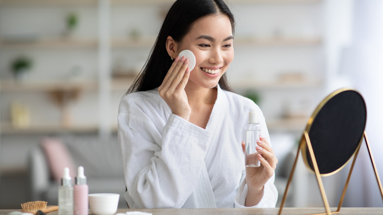 Woman applying toner with cotton pad onto face and looking into mirror