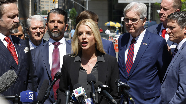 Pam Bondi addressing the media in a black blazer and dark grey blouse