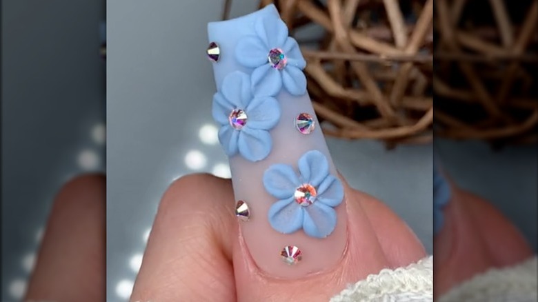 Close-up of 3D nail art, with a blue and pink ombre base, painted flowers, and rhinestones