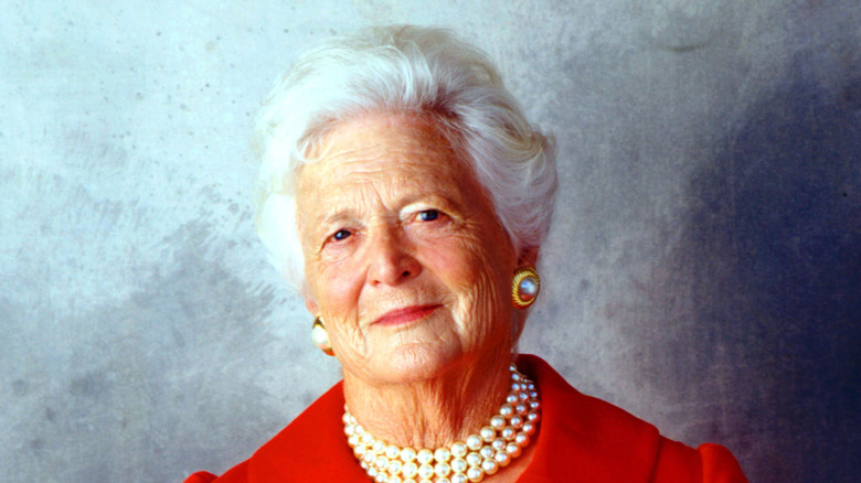 Barbara Bush smirking in a photograph while wearing a red dress and red lipstick.