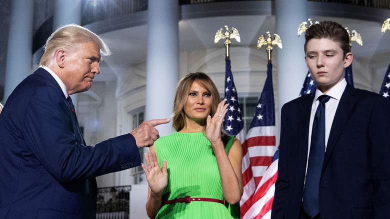 Barron Trump looking glum in a suit while his father Donald Trump points at him and mom Melania waves