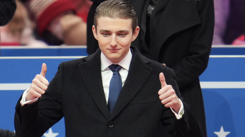 Barron Trump giving a thumbs-up to a crowd in a suit with slicked-back hair