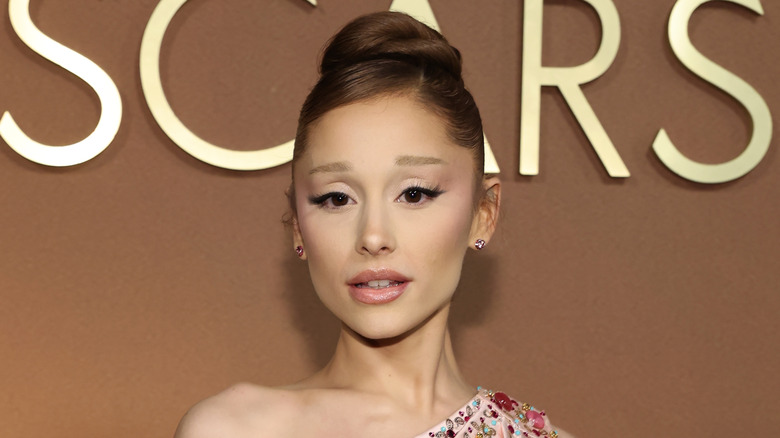 Ariana Grande in pink dress with hair in bun in front of Oscars sign.