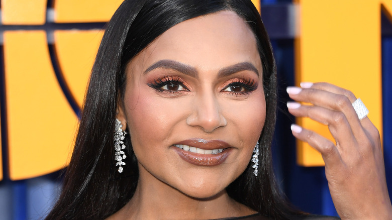 Mindy Kaling smiling and holding a hand up beside her face.