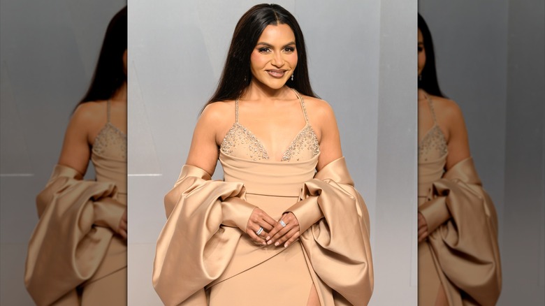 Mindy Kaling posing in a beige spaghetti strap gown with detached sleeves and crystal detailing on the bust.
