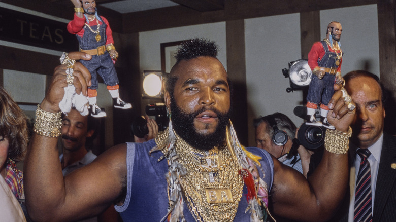 Mr. T, wearing a blue t-shirt, with several necklaces, bracelets and rings, poses with his action figures.