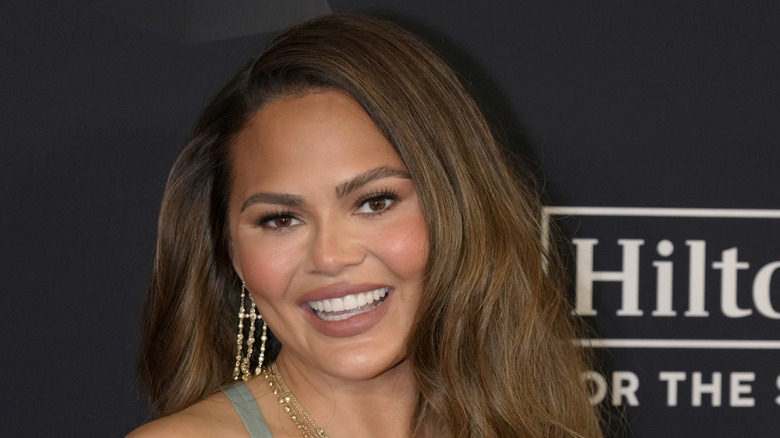 Chrissy Teigen smiling on a Hilton sponsored red carpet in a sage green dress with gold jewelry