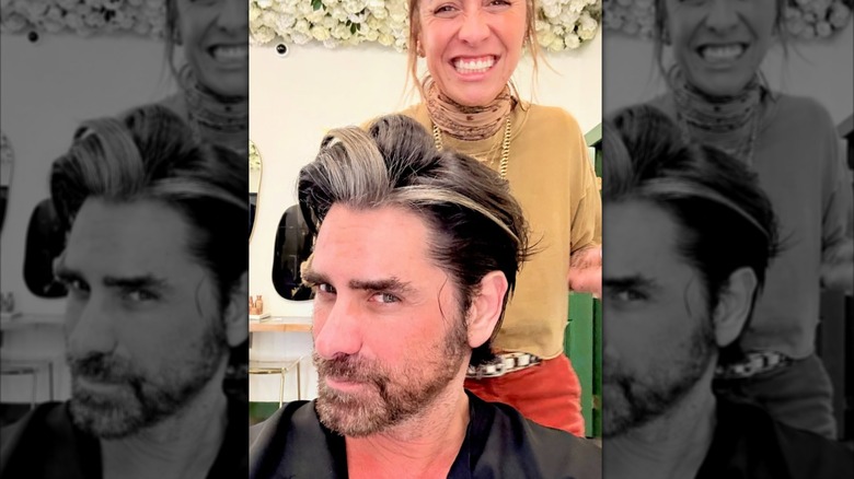 John Stamos taking a selfie of the blond streaks in his hair with his hairdresser smiling behind him
