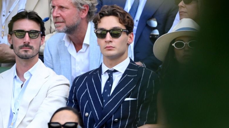 George Russell watching tennis at Wimbledon in a pinstripe suit and wearing sunglasses.