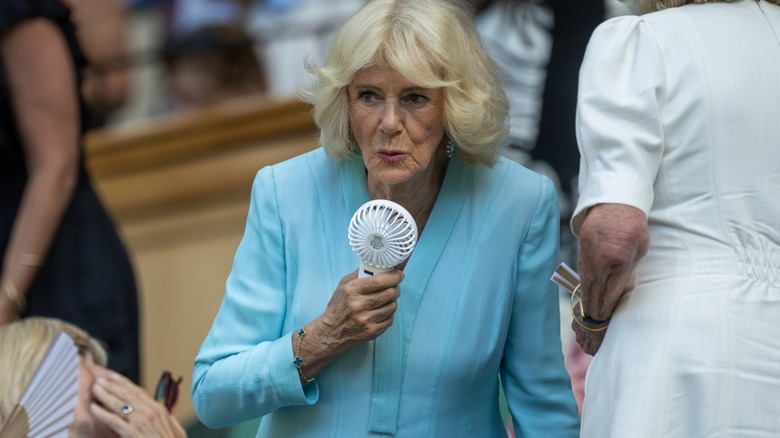 Queen Camilla using a portable fan and wearing a turquoise dress at Wimbledon 2026.