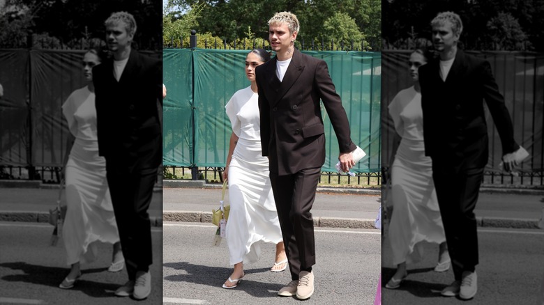 Romeo Beckham in a suit and girlfriend Kim Turnbull in a white dress walk across the street in Wimbledon.