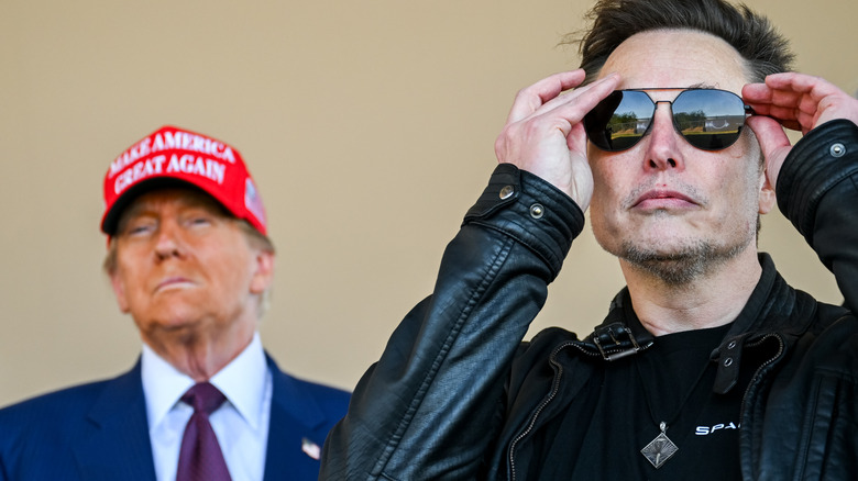 Elon Musk adjusting his sunglasses with Donald Trump standing behind him
