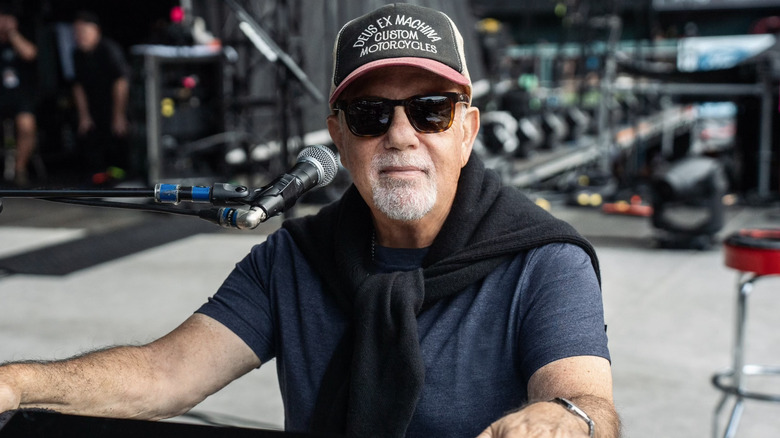 Billy Joel sitting in front of a microphone wearing a hat and sunglasses
