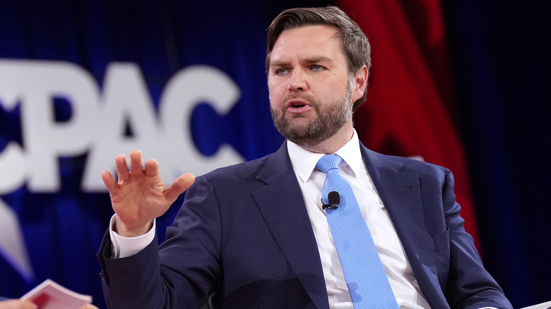JD Vance talking while raising his hand in a blue suit