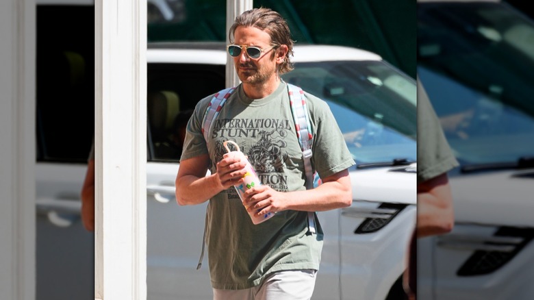 Bradley Cooper walking around New York with a green shirt, shades, a backpack, and a juice bottle.