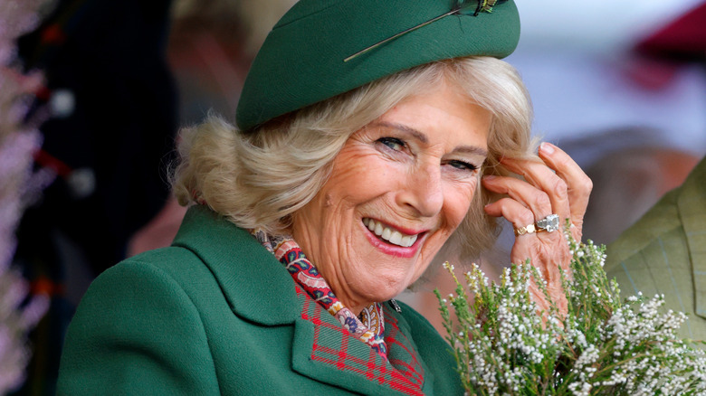 Queen Camilla smiles while wearing a green hat and suit and holding flowers
